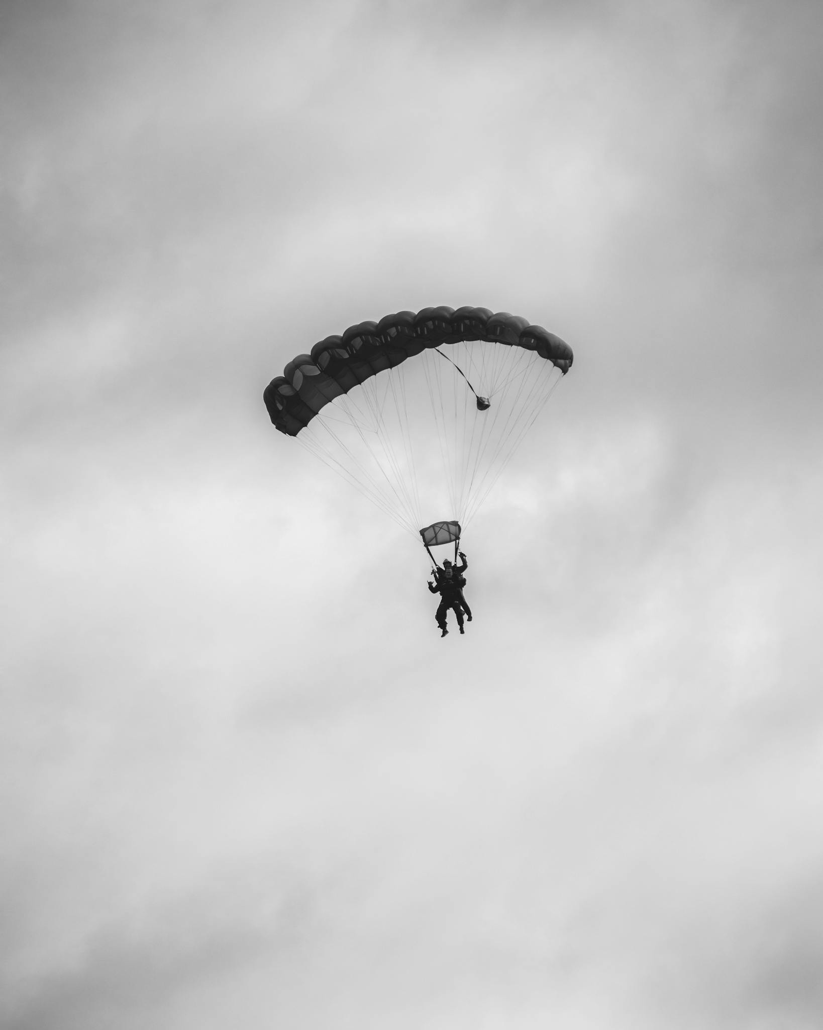 Silhouette of two people tandem skydiving with parachute against a cloudy sky.